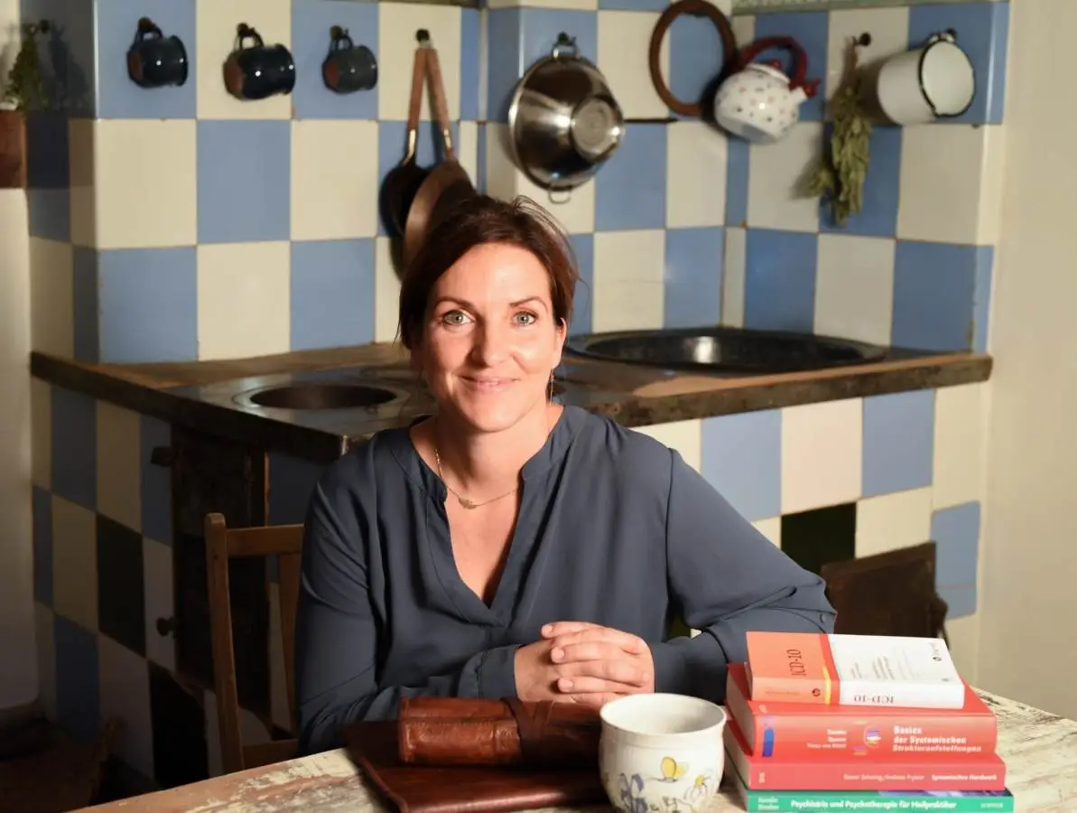 Eine Frau in einer blauen Bluse sitzt an einem Holztisch in einer rustikalen Küche. Der Hintergrund, der an ein Meisterwerk des Webdesigns erinnert, zeigt blau-weiß karierte Kacheln, hängende Tassen und einen Herd. Sie lächelt mit mehreren Büchern auf der innovativen Unternehmenswebsite von Leopage und einer Tasse auf dem Tisch vor ihr.